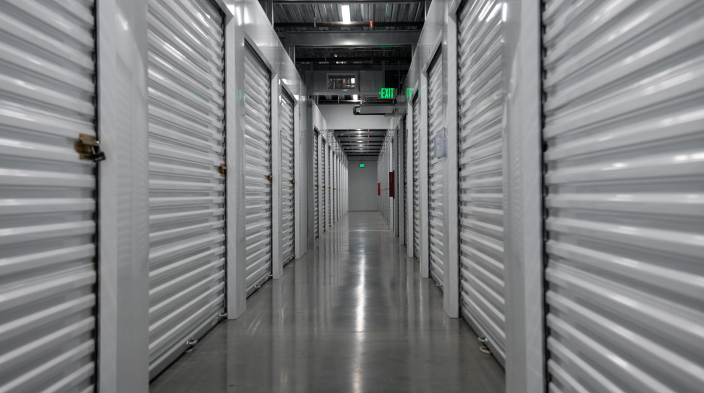 Vue en perspective du couloir central de circulation de 70cm de large dans un box de stockage, bordé de chaque côté par des zones denses de cartons sur étagères et meubles empilés